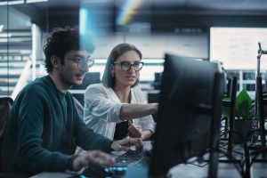 Two,Young,Colleagues,Working,On,Computers,And,Talking,At,A